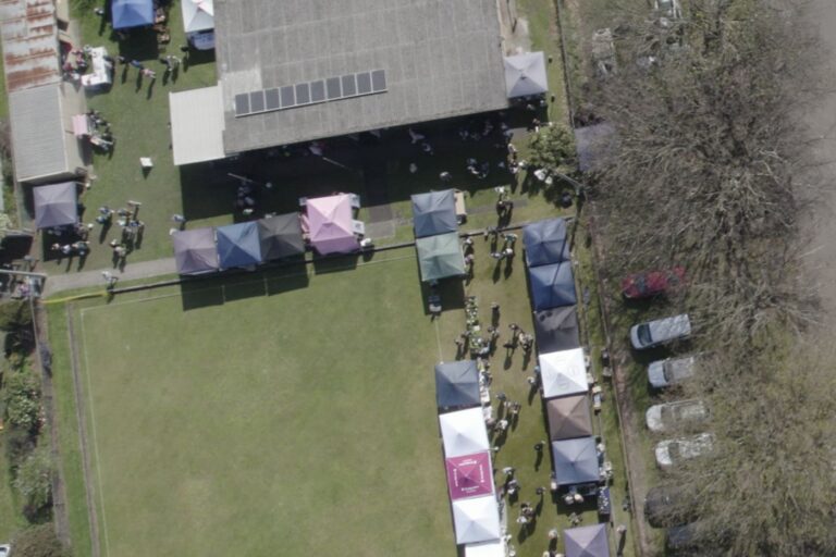 Market day at the Hub from directly above, stall canopies lining two sides of the green with shoppers between them