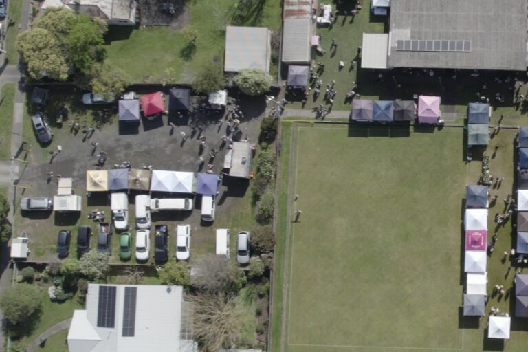 An aerial view of market day showing stalls around the green, the carpark full of cars and vans, and people throughout