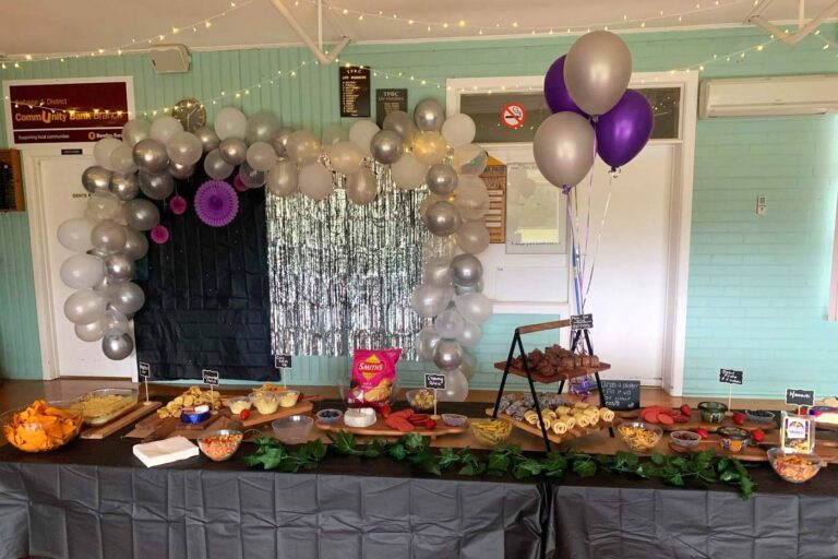 The hall dressed for a party, with a silver and purple balloon arch, fairy lights and a long table of food