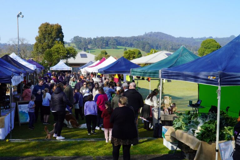 Shoppers crowd the grass between two rows of blue, white and green marquees at the Trafalgar Farmers Market, with green hills behind
